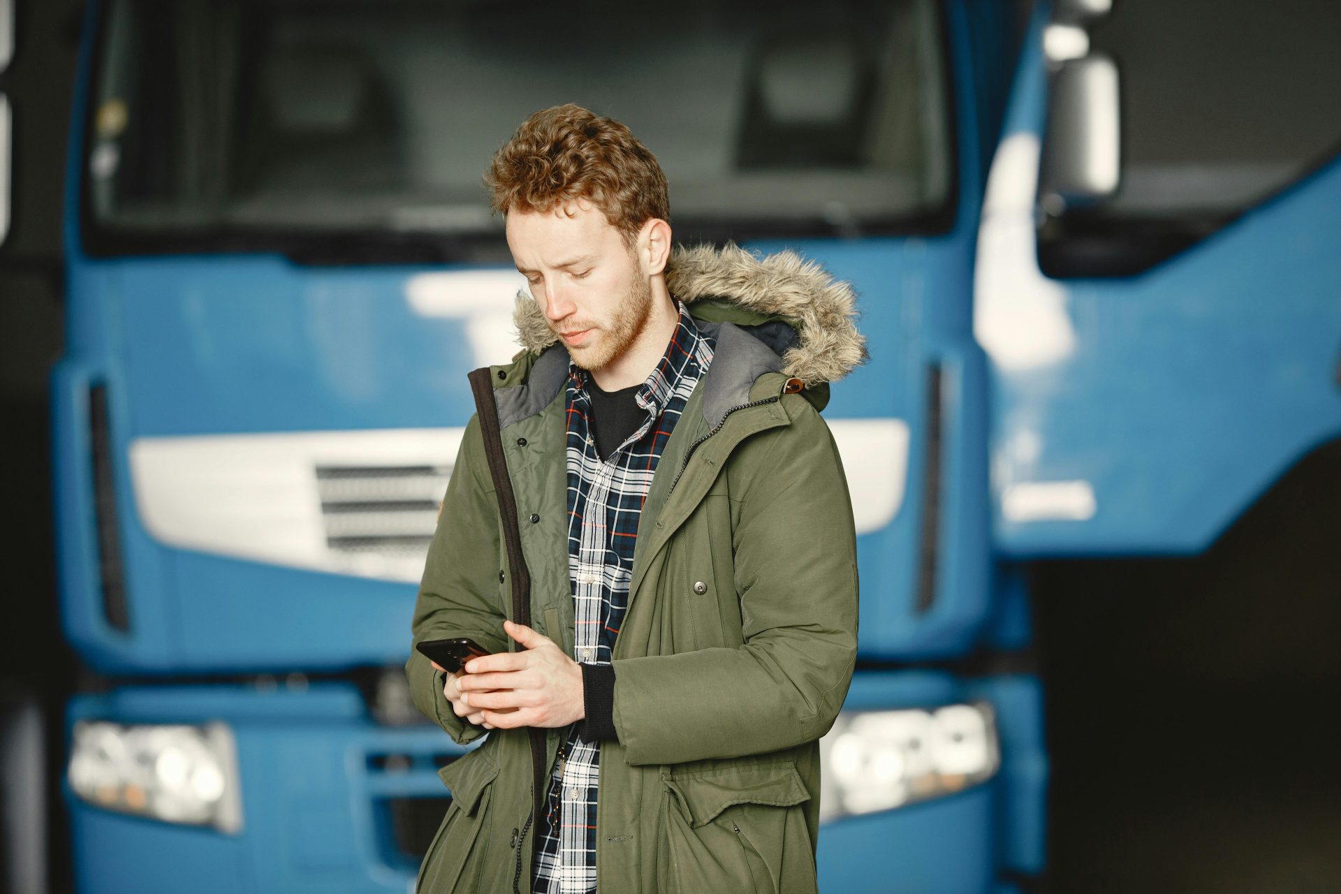 A man wearing a parka checks his mobile phone in front of a blue truck parked in a garage.